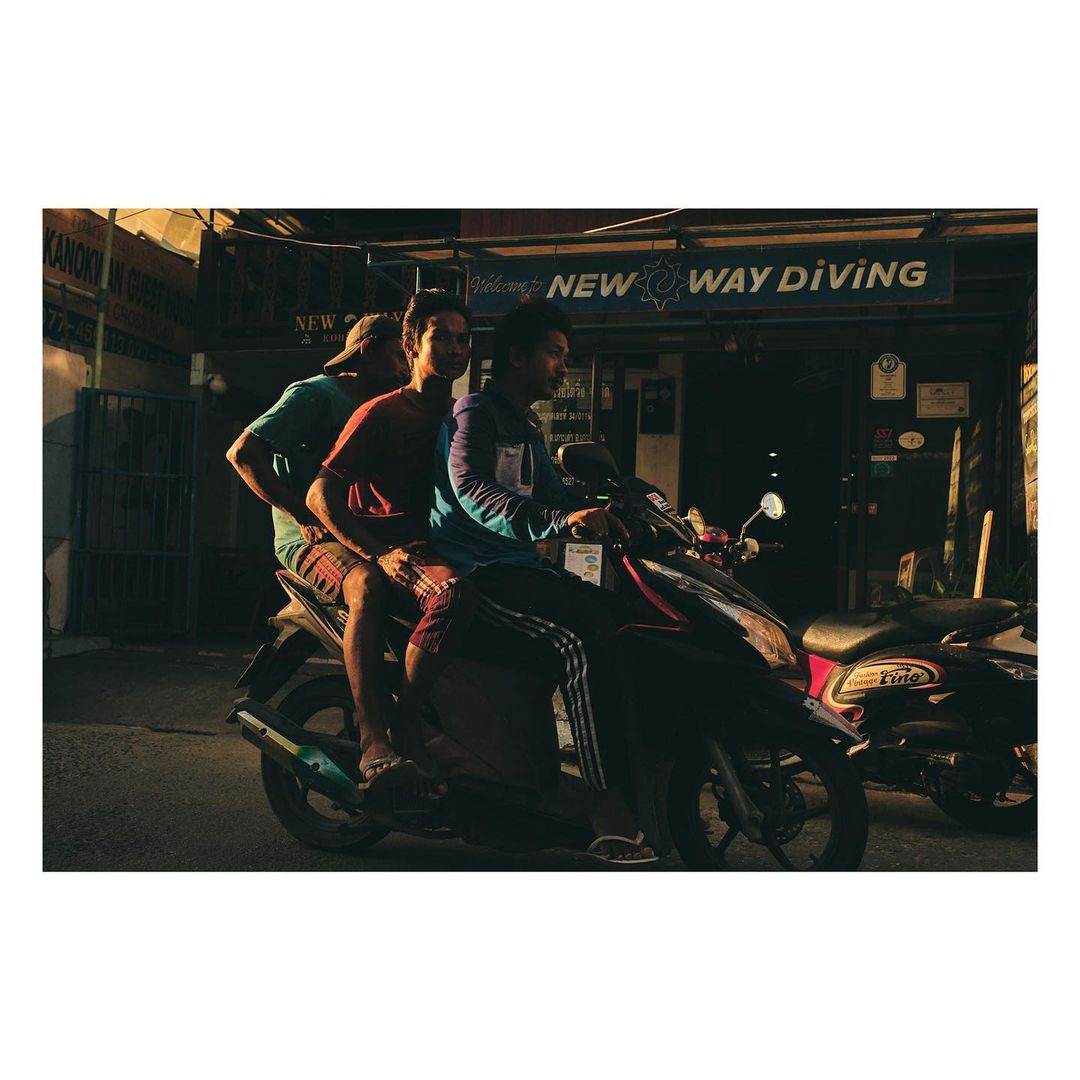 Three adult males ride a black motorcycle on a street near a building with 'NEW WAY DIVING' signage during daytime, captured in a medium shot.