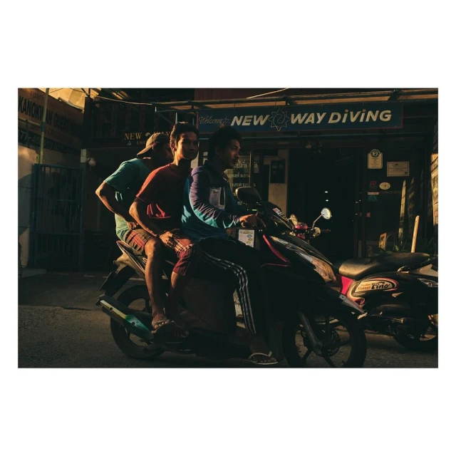 Three adult males ride a black motorcycle on a street near a building with 'NEW WAY DIVING' signage during daytime, captured in a medium shot.