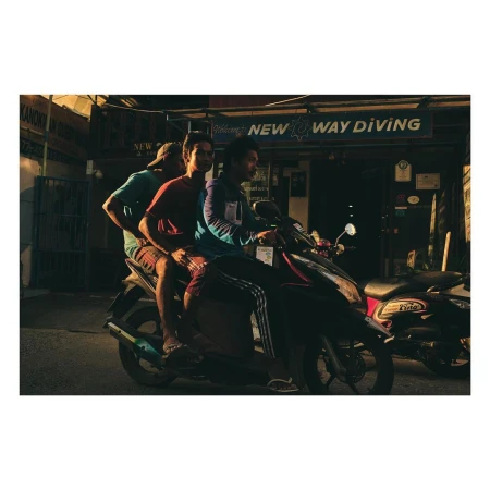 Three adult males ride a black motorcycle on a street near a building with 'NEW WAY DIVING' signage during daytime, captured in a medium shot.