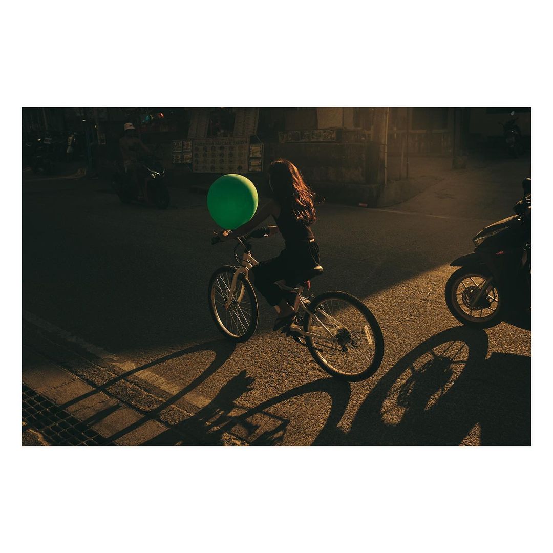 Young woman riding a white bicycle in a street with a green balloon, lit by golden sunlight, casting shadows on the asphalt. Motorbikes nearby.