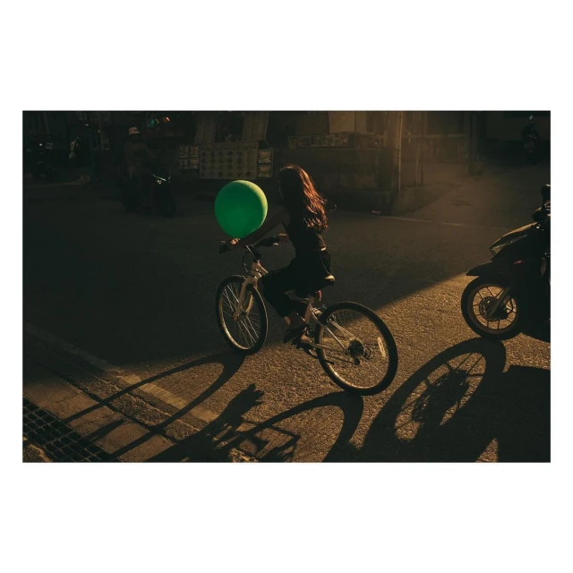 Young woman riding a white bicycle in a street with a green balloon, lit by golden sunlight, casting shadows on the asphalt. Motorbikes nearby.