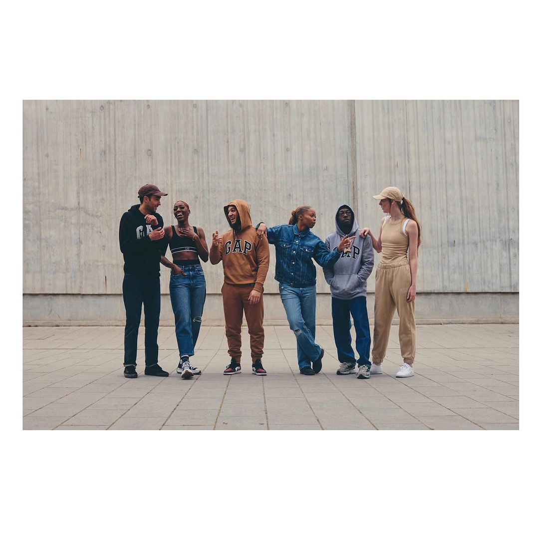 Group of six diverse young adult models in casual clothing by GAP standing in front of a concrete wall, posing in an outdoor setting.