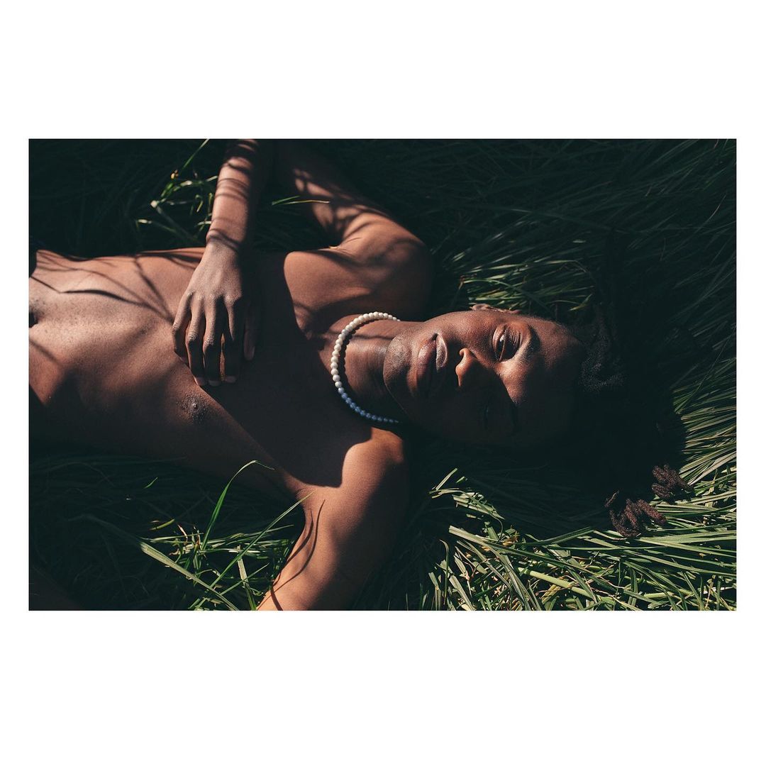 Shirtless Black man wearing a pearl necklace lying in tall green grass outdoors in bright sunlight.
