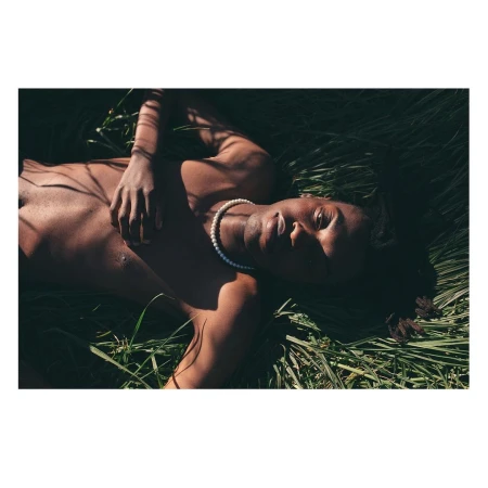 Shirtless Black man wearing a pearl necklace lying in tall green grass outdoors in bright sunlight.