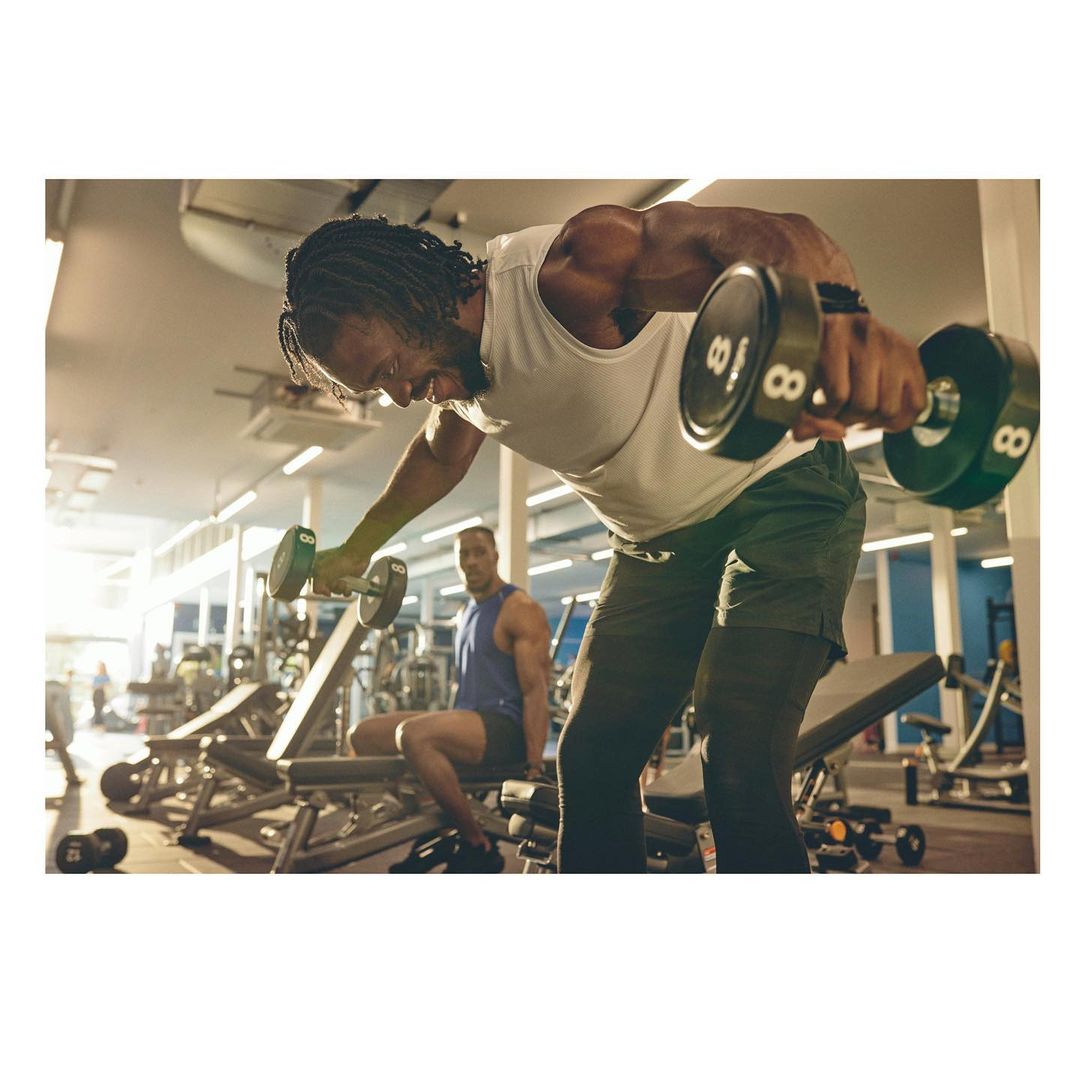 Muscular man lifting dumbbells at the gym, performing a lateral raise. Another man sits on a weight bench in the background.