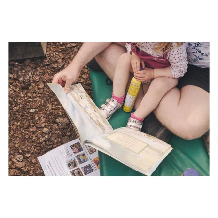 Child and adult seated outdoors, reading an illustrated book on a green bench. Sunscreen nearby, surrounded by mulch.