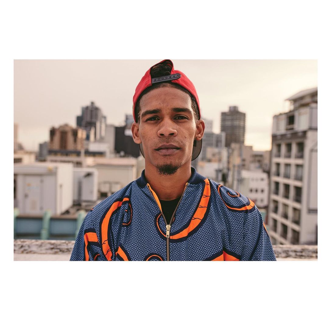 Waist-up portrait of a young adult man in a patterned jacket and backwards red cap, set against an urban rooftop and cityscape backdrop.