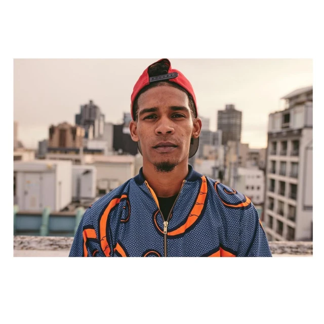 Waist-up portrait of a young adult man in a patterned jacket and backwards red cap, set against an urban rooftop and cityscape backdrop.