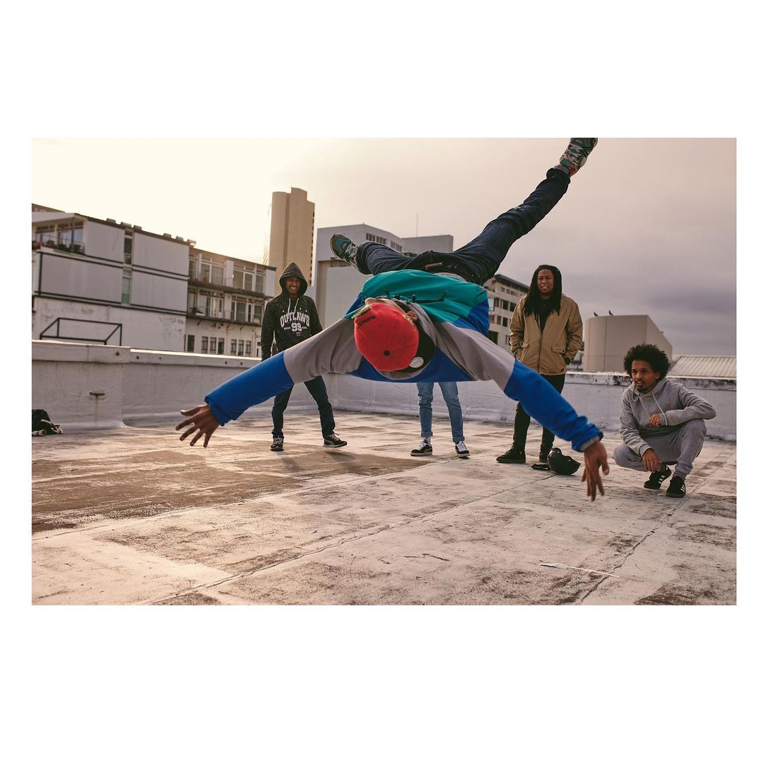 Acrobatic man in mid-air performing a handstand on a rooftop with four other people watching in the background on a sunny day.