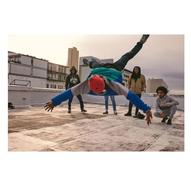 Acrobatic man in mid-air performing a handstand on a rooftop with four other people watching in the background on a sunny day.