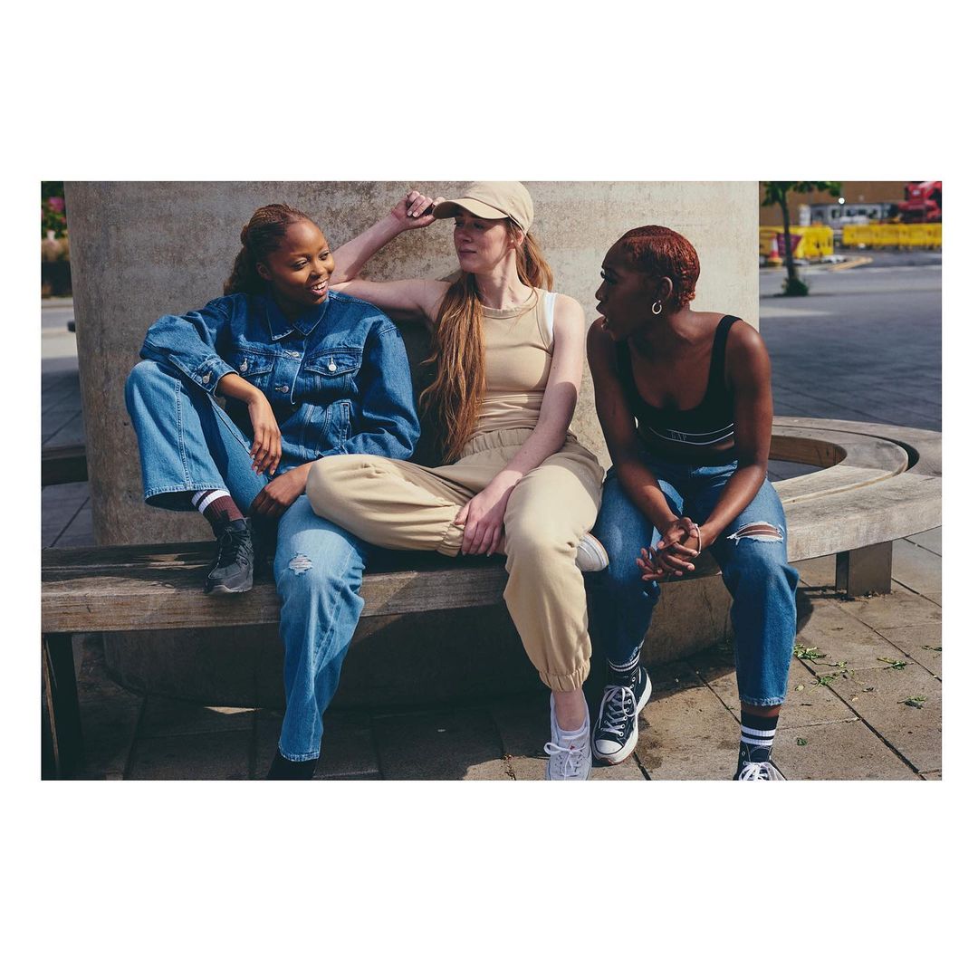 Three women in casual outfits are sitting on a concrete bench outdoors, engaged in conversation on a sunny day in an urban environment.