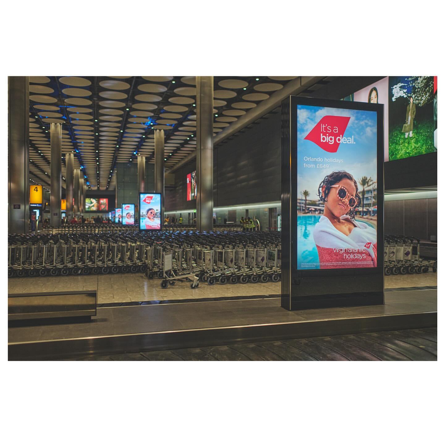 Airport terminal interior showcasing luggage carts and a Virgin Holidays advertisement featuring a woman in sunglasses by a pool.