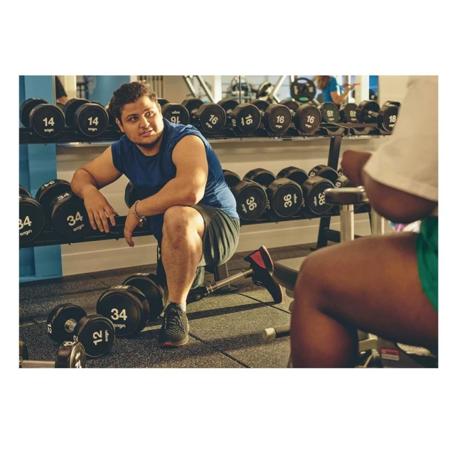 Man resting at the gym after workout, seated with dumbbells and weights in the background, fitness and exercise setting, indoor shot.