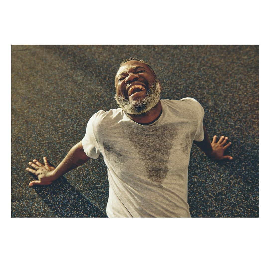 Sweaty adult man with a grey beard lying on his back on a grey floor, wearing a white t-shirt, seen from a high angle with shallow depth of field.