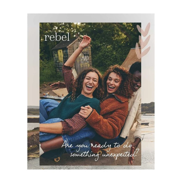 Three cheerful young women enjoy time together outdoors, posing on a skateboard against a backdrop of greenery and a slogan, promoting the 'rebel' brand.