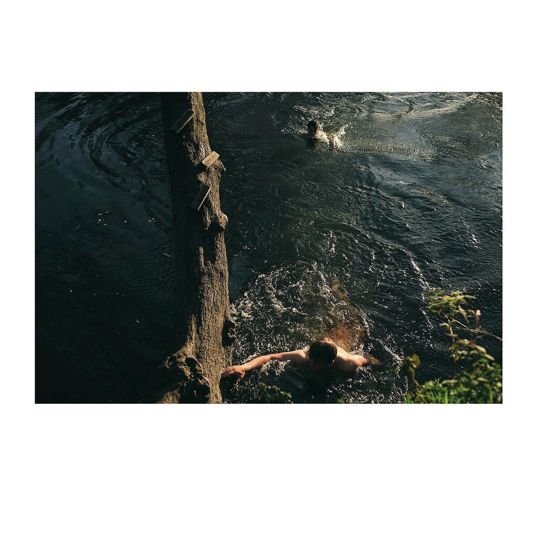 Two people swimming in a dark river surrounded by trees on a sunny day. One person is in the foreground; the other is farther away.