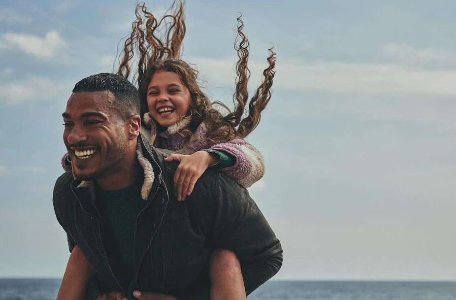 A smiling dark-skinned adult carries a laughing child on his back near a body of water under a bright, partly cloudy sky.