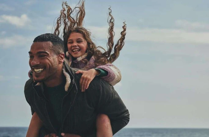 A smiling dark-skinned adult carries a laughing child on his back near a body of water under a bright, partly cloudy sky.