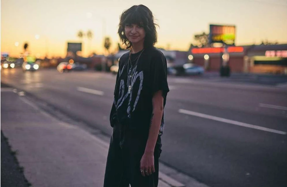 Young adult with dark hair stands on a city street at dusk, wearing a black t-shirt and pants, with blurred traffic and buildings.