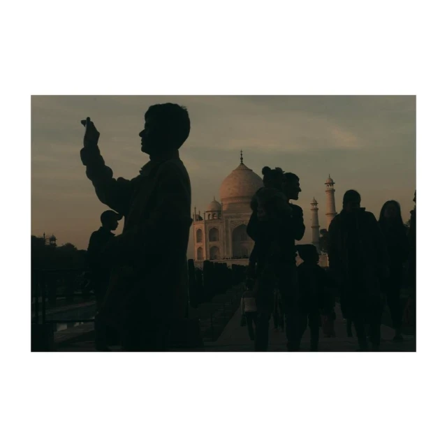 Silhouetted tourists visiting the Taj Mahal at dusk. The marble mausoleum is visible in the background with a cloudy sky.