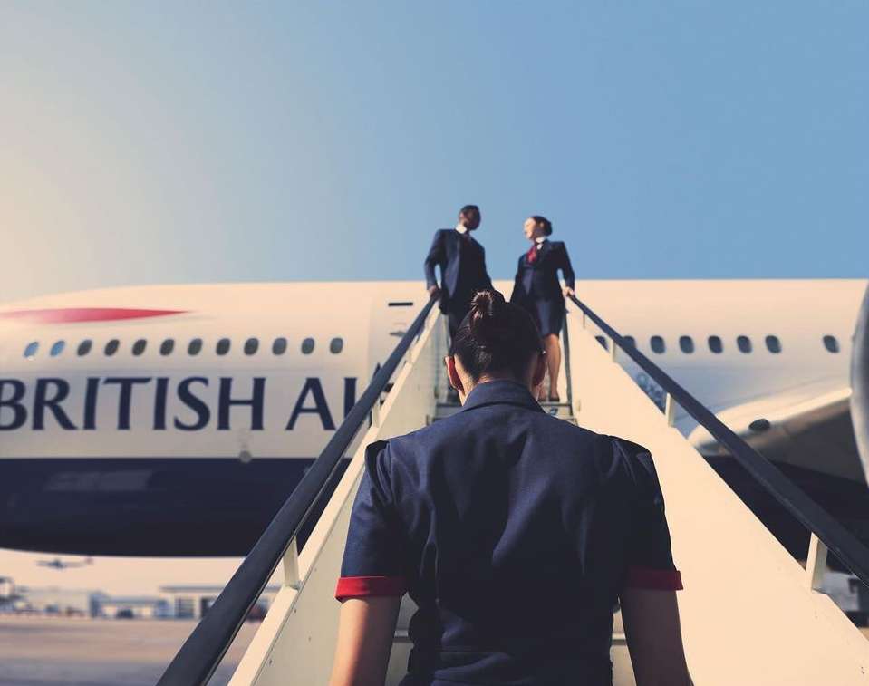 Airplane boarding staircase with flight attendants in navy blue uniforms, one African American male, one Caucasian female, and the tail of the airplane.