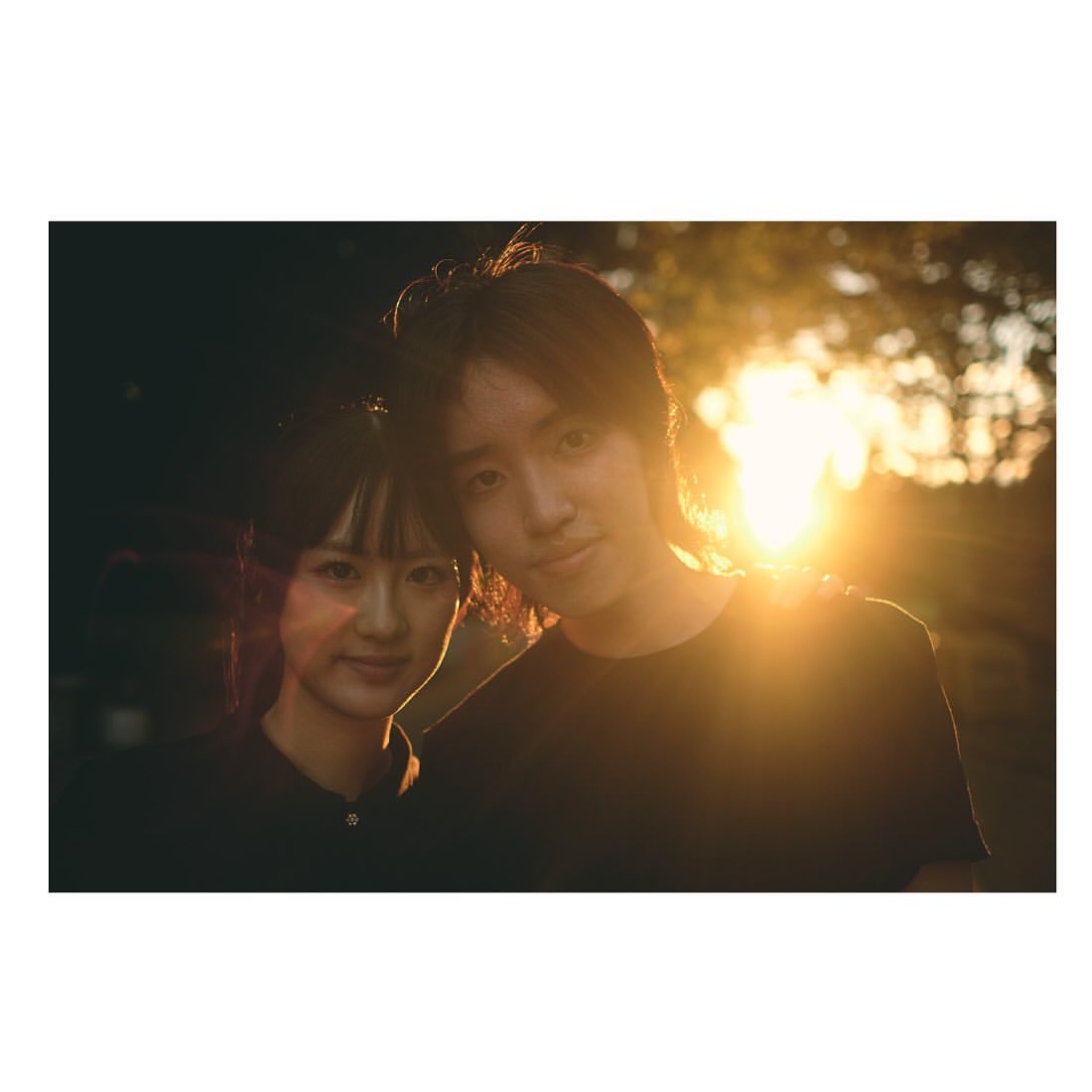 Portrait of a smiling young couple in an outdoor setting during golden hour, backlit by the sun, creating a warm, romantic atmosphere.