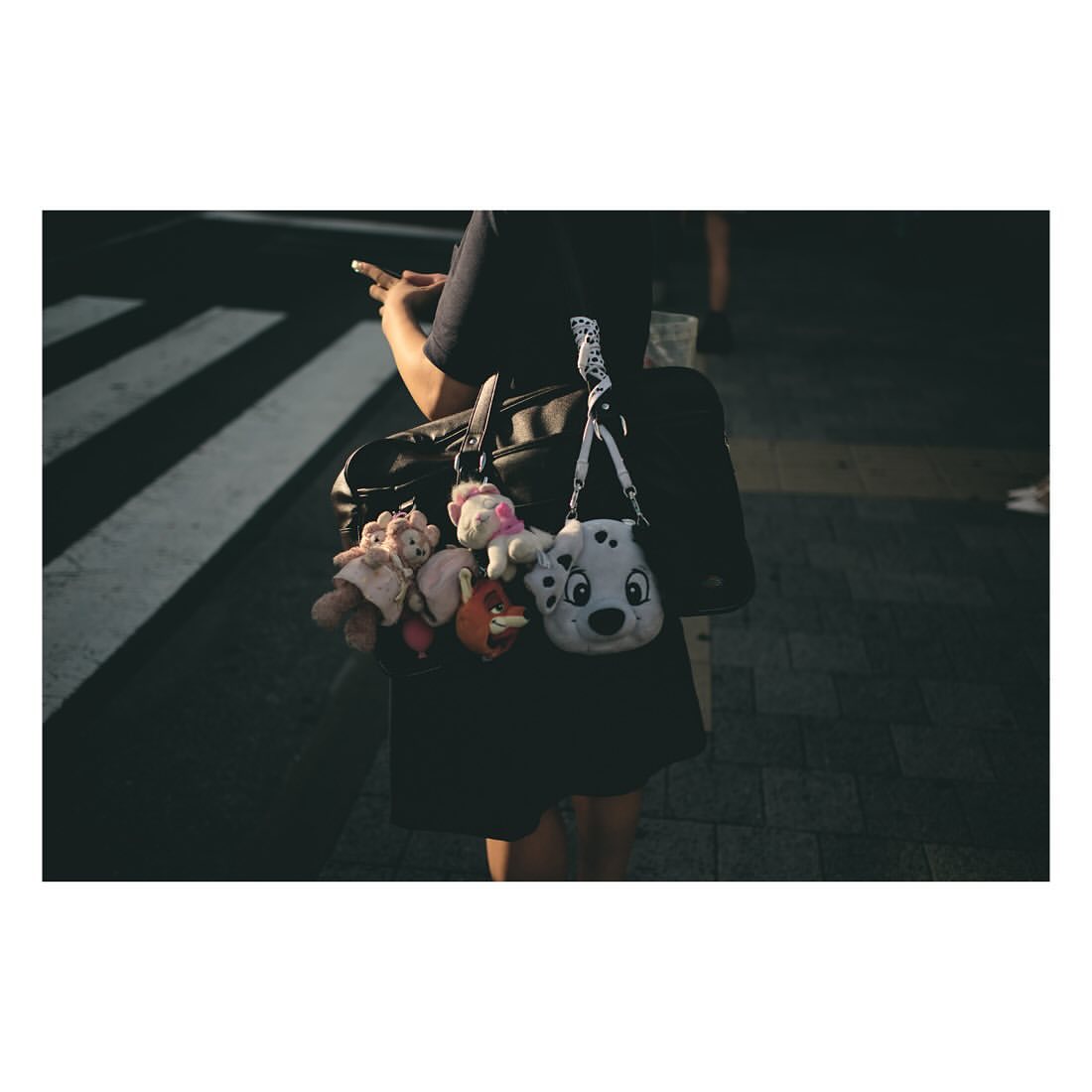 A person wearing a black dress holds a black bag with several plush toys hanging from it while crossing a zebra-striped crosswalk.