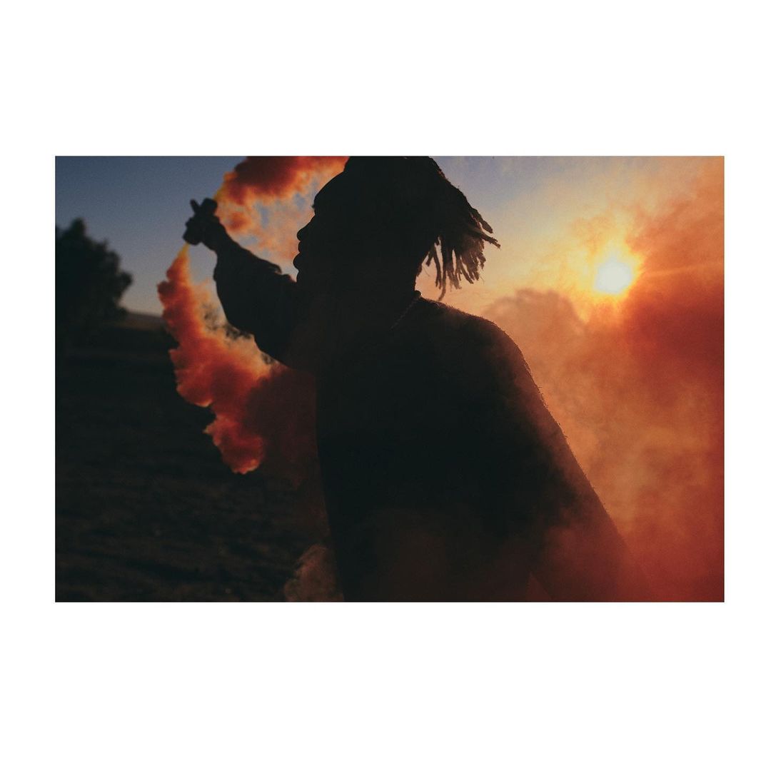 Silhouette of a person with dreadlocks holding an orange smoke grenade in a landscape against the sunset with trees in the background.