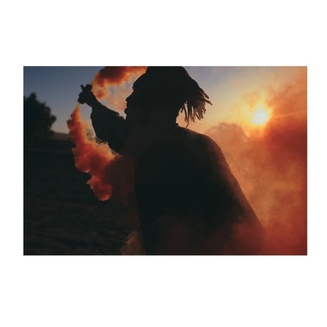 Silhouette of a person with dreadlocks holding an orange smoke grenade in a landscape against the sunset with trees in the background.