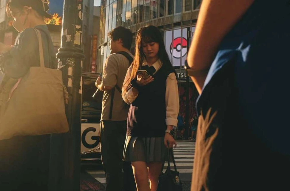 An Asian teenage girl looks at her mobile phone standing on a city street while pedestrians walk by in this candid outdoor shot.