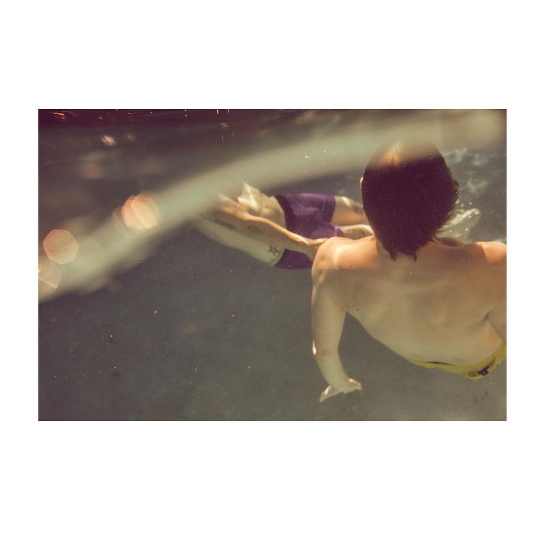 Two individuals are underwater, one in motion and the other with their back to the camera, swimming in a blurry, muted light.