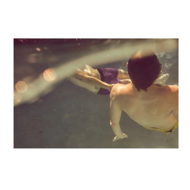 Two individuals are underwater, one in motion and the other with their back to the camera, swimming in a blurry, muted light.
