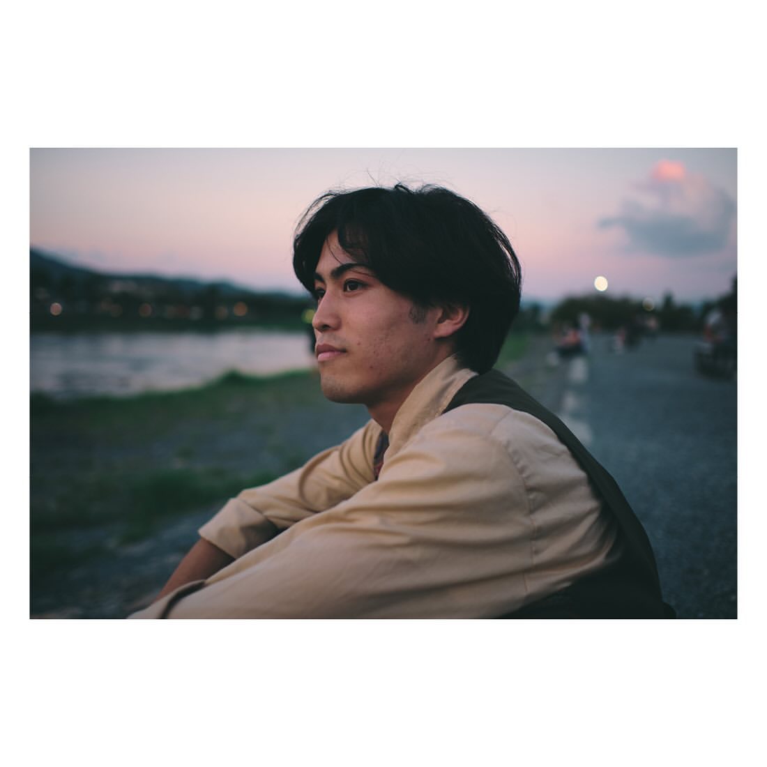 Young man in casual clothes seated near a river at sunset. Soft, warm lighting creates a contemplative mood in an outdoor setting.
