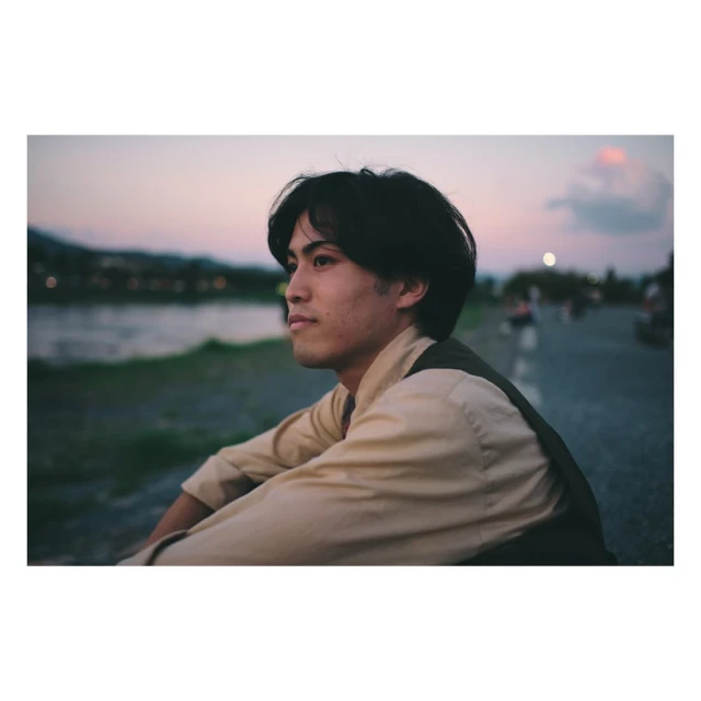 Young man in casual clothes seated near a river at sunset. Soft, warm lighting creates a contemplative mood in an outdoor setting.