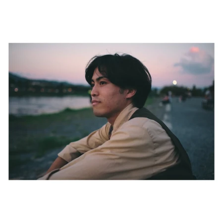 Young man in casual clothes seated near a river at sunset. Soft, warm lighting creates a contemplative mood in an outdoor setting.