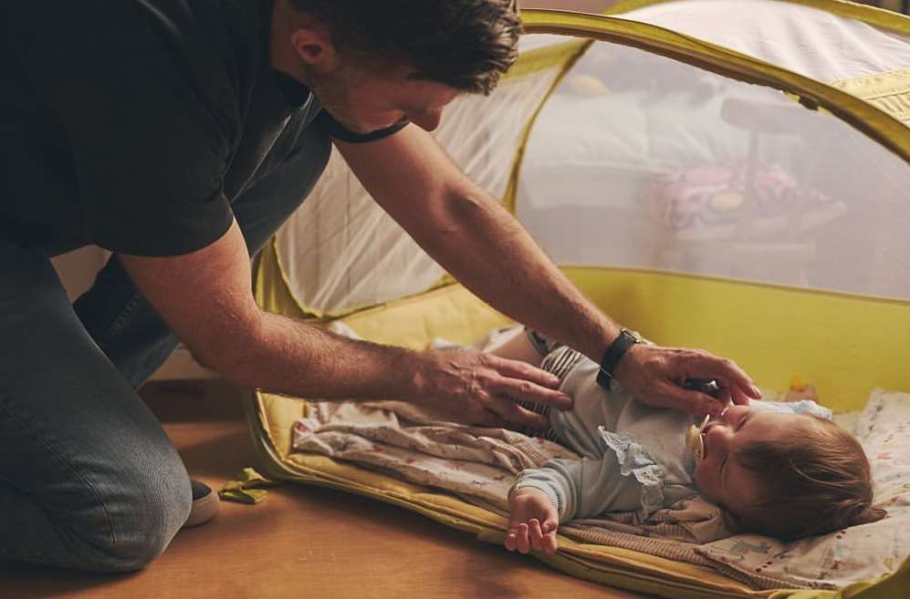 Baby lying in a portable crib with an adult male leaning over, gently touching the baby. Indoor setting with warm lighting.