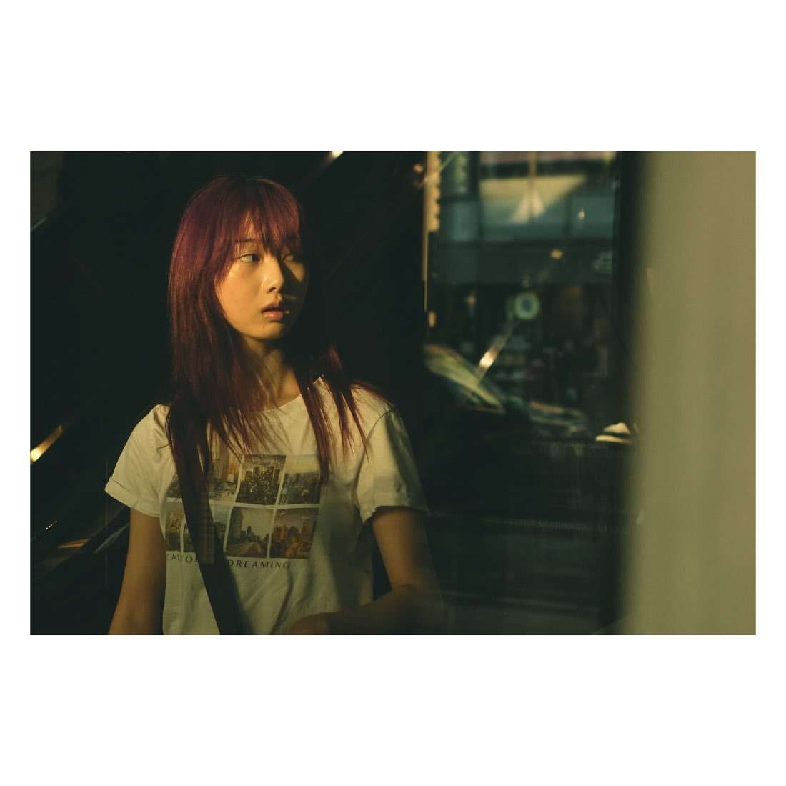 Young woman with auburn hair stands indoors with a white t-shirt, a purse strap, and a vehicle visible through the glass in the background.