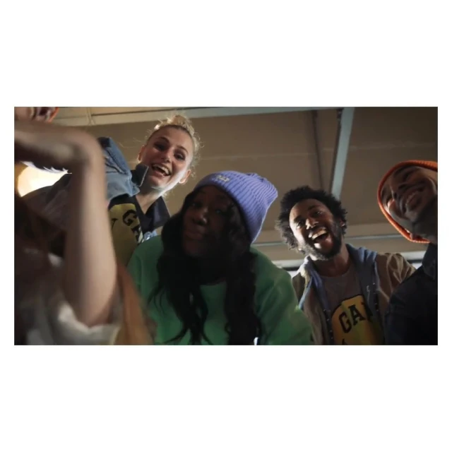 Diverse group of smiling people looking down at the camera, wearing casual clothing, including hats and a Gap-branded shirt, indoors.