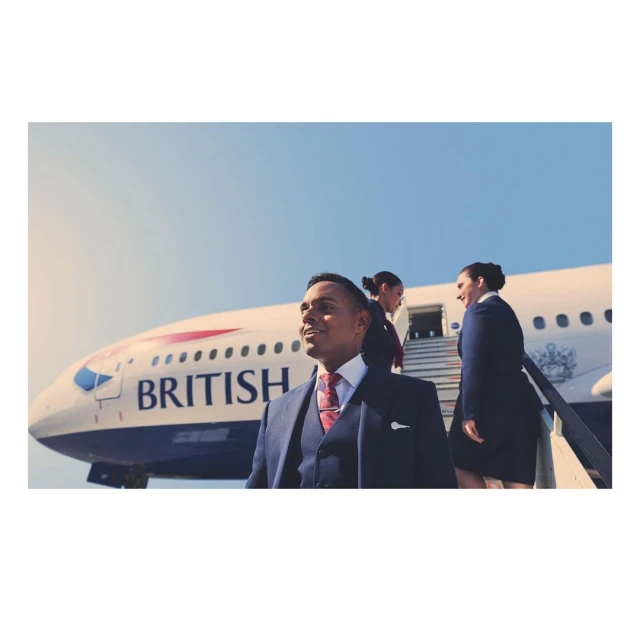 Male airline pilot in uniform poses with flight attendants on airplane steps under clear blue sky with British Airways plane visible.