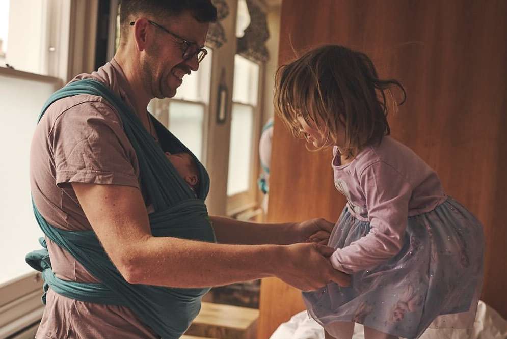 Father holding a newborn baby in a wrap while holding hands with his young daughter, inside a home with natural lighting.