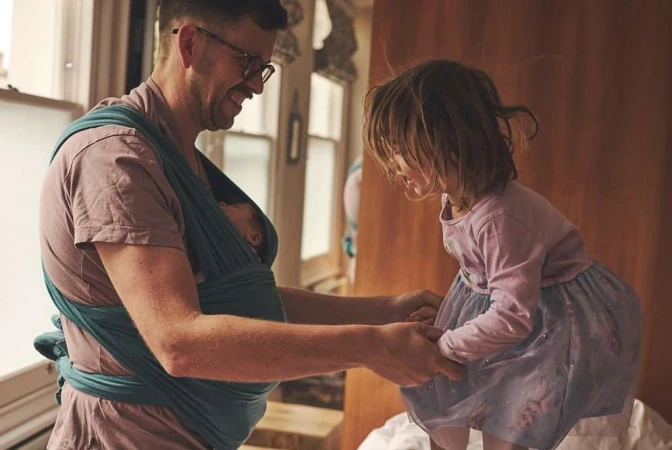 Father holding a newborn baby in a wrap while holding hands with his young daughter, inside a home with natural lighting.