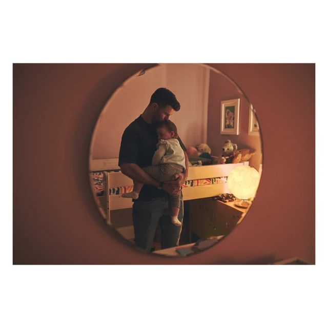 A man holds a sleeping baby in a nursery as seen in an oval mirror with soft lighting, creating a heartwarming family moment.