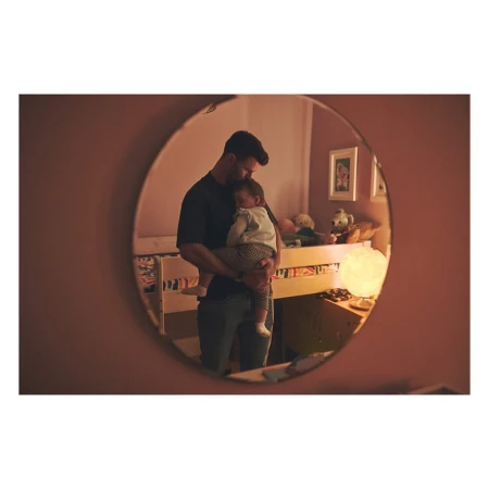 A man holds a sleeping baby in a nursery as seen in an oval mirror with soft lighting, creating a heartwarming family moment.