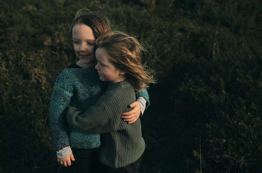 Two young children embrace outdoors in a field, wearing knitted sweaters, with soft, diffused lighting and a natural, grassy background.