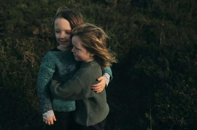 Two young children embrace outdoors in a field, wearing knitted sweaters, with soft, diffused lighting and a natural, grassy background.