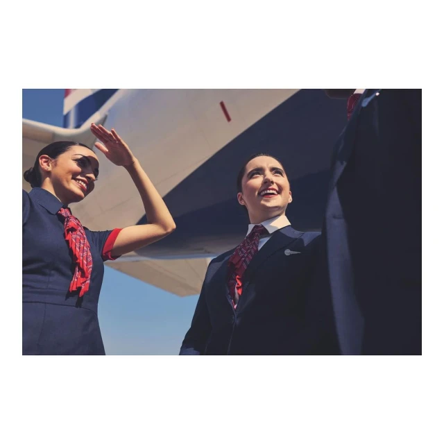 Two smiling flight attendants in uniform stand outside an airplane on a sunny day, one waving, showcasing air travel and service.