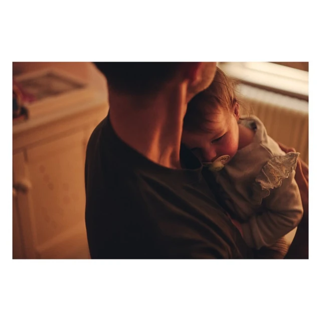 Father cradling sleeping newborn baby indoors in warm lighting. The baby has a pacifier in mouth, and the father wears a dark green t-shirt.