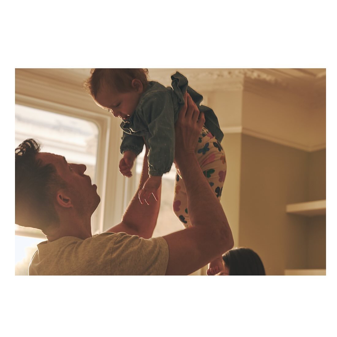 Father joyfully lifts his baby daughter into the air indoors by a window in warm light, showcasing a tender family moment.