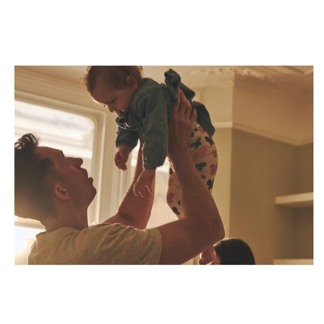 Father joyfully lifts his baby daughter into the air indoors by a window in warm light, showcasing a tender family moment.