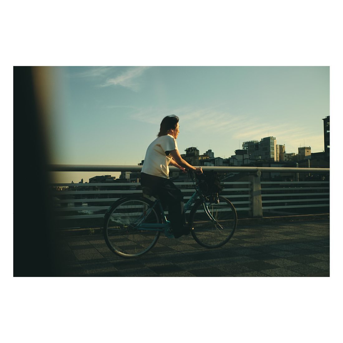 Side view of an adult riding a bicycle on a bridge in the city. The person wears a casual t-shirt and the sky is blue in the background.